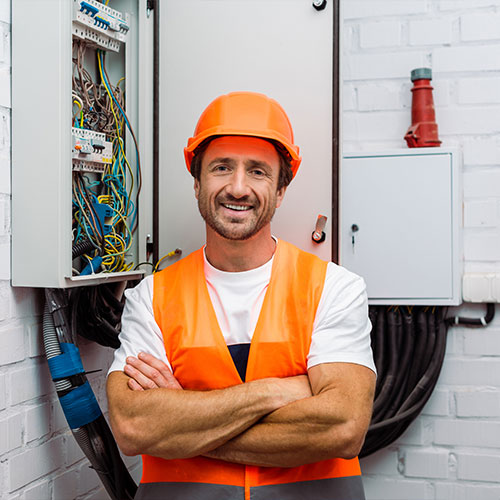 Handsome electrician with crossed arms smiling at camera near electric panel